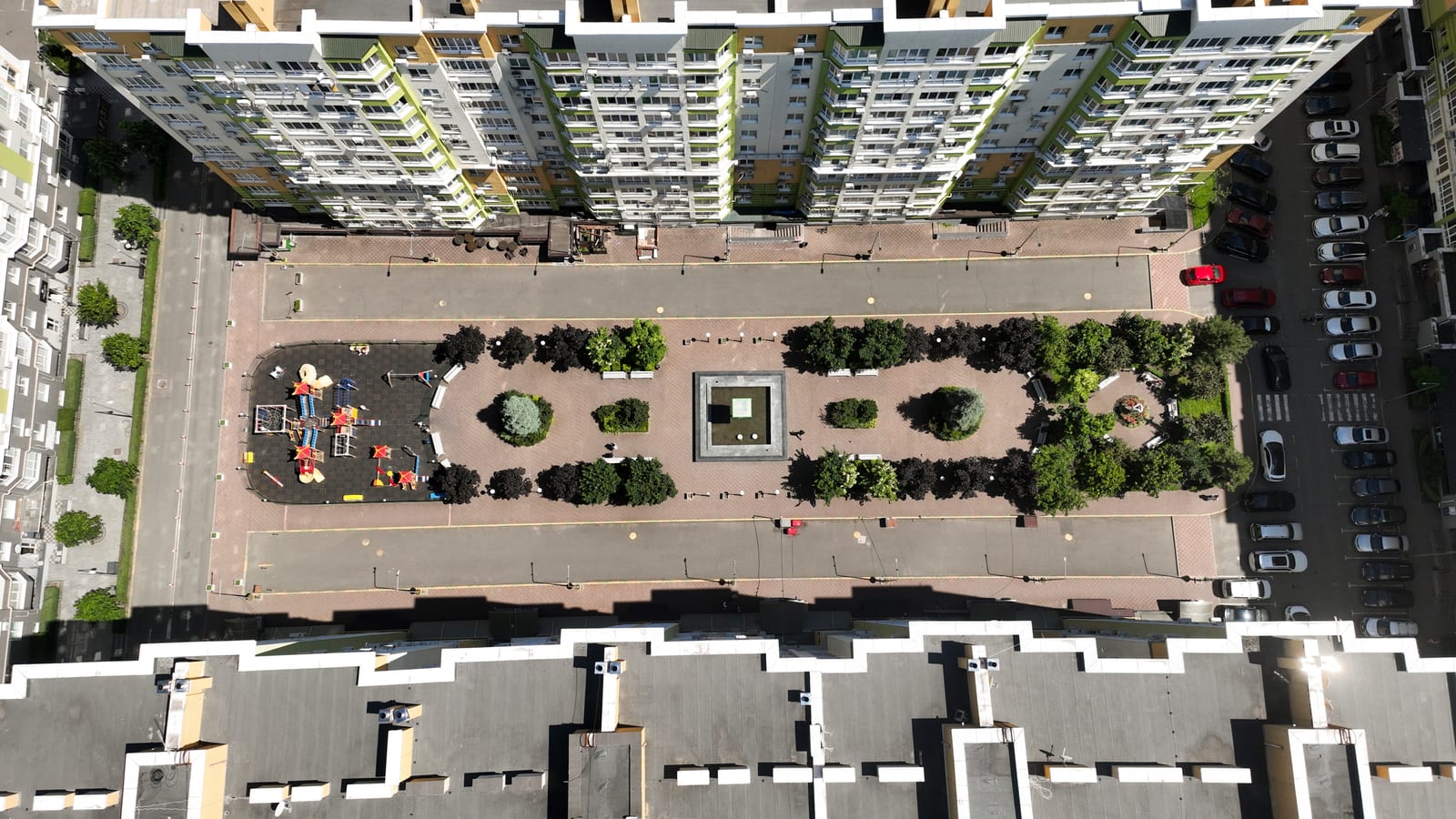 A long planted boulevard between two rows of SAVO blocks seen straight from above, with a formal strip of planting down the middle and a playground at one end