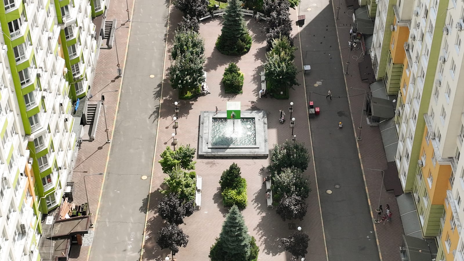 The boulevard between two blocks of Evromisto by SAVO seen from above: a working fountain, clipped trees, benches and residents walking