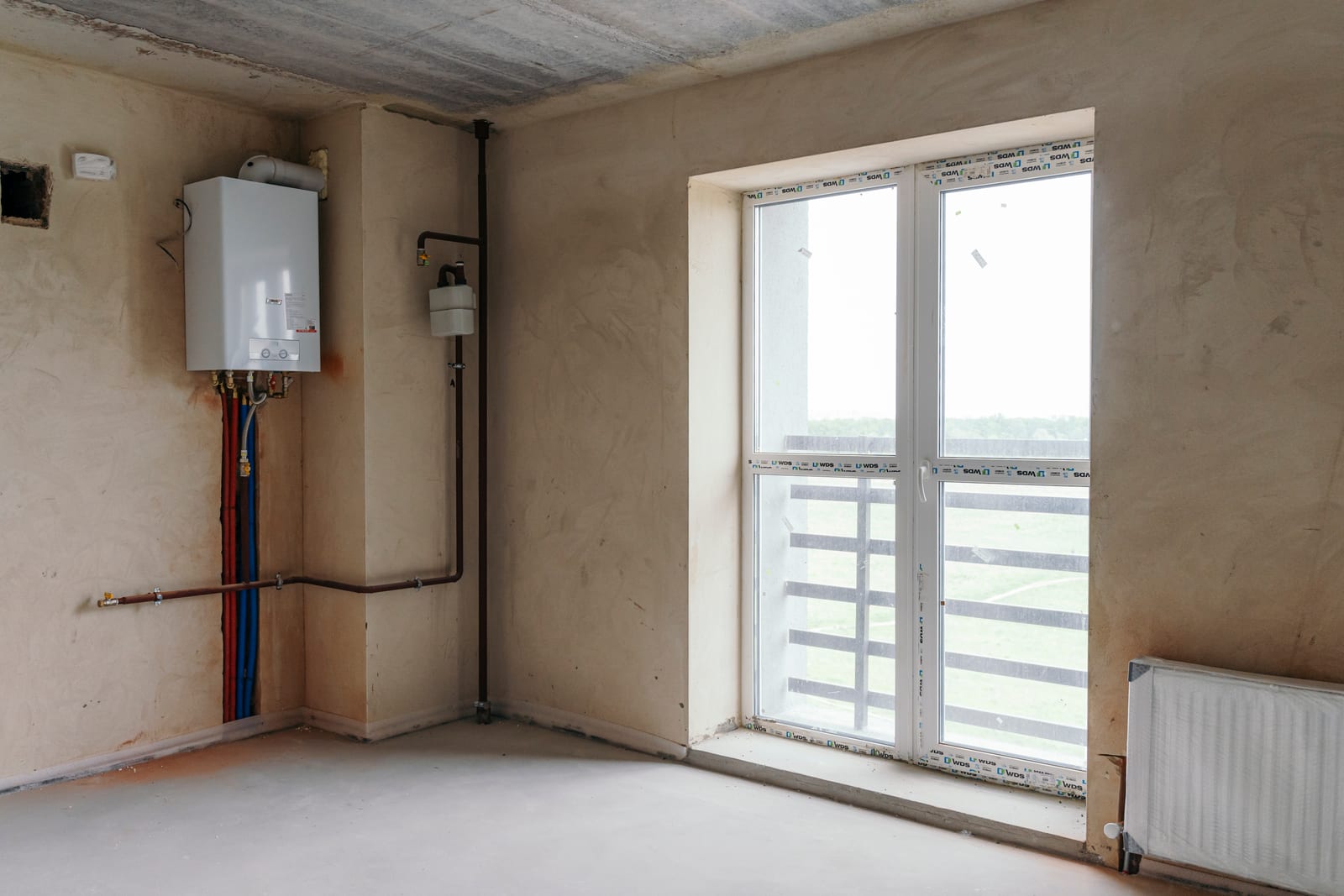 A room handed over unfinished: screed, plaster, a radiator, the boiler and a window onto open fields