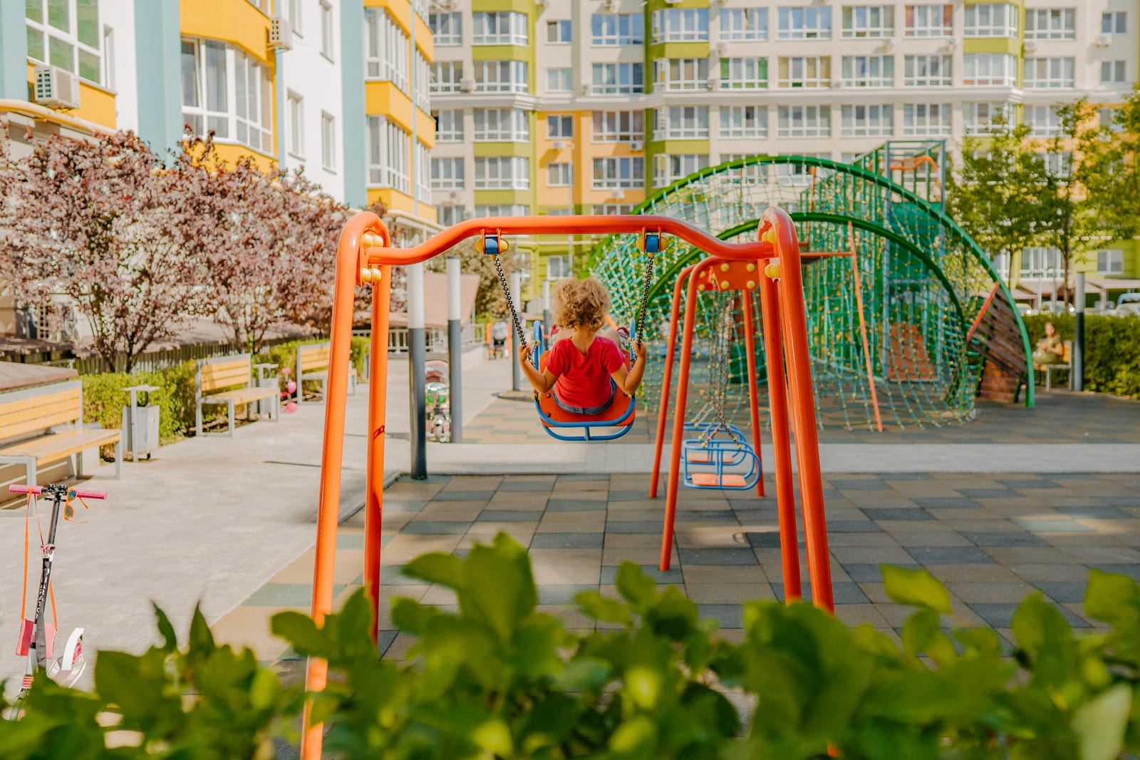 A child in red on an orange-and-green swing beneath a pink magnolia in full flower, in a SAVO courtyard.