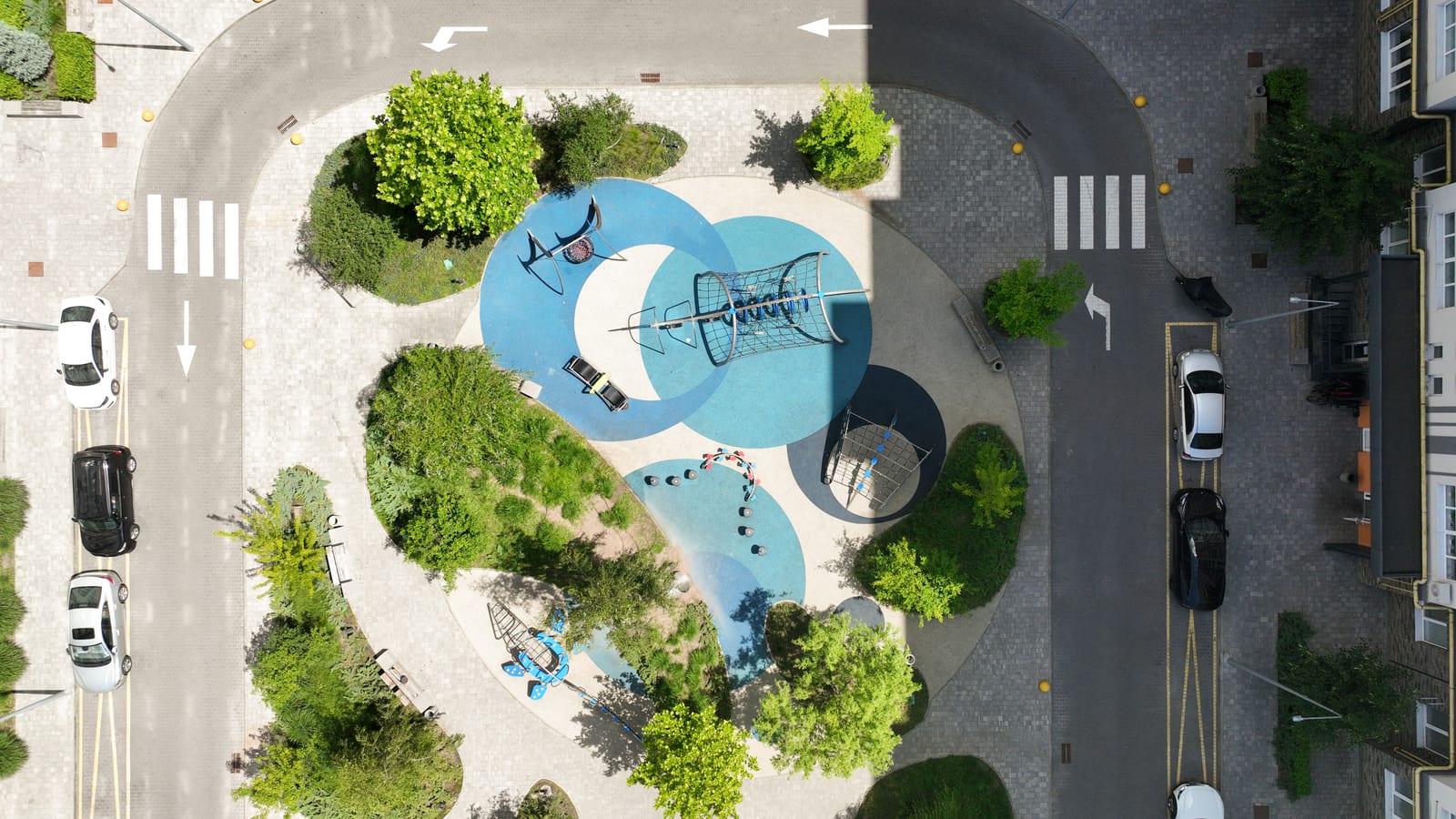 A playground straight from above: turquoise safety surfacing, climbing frames on it and trees around its edge.
