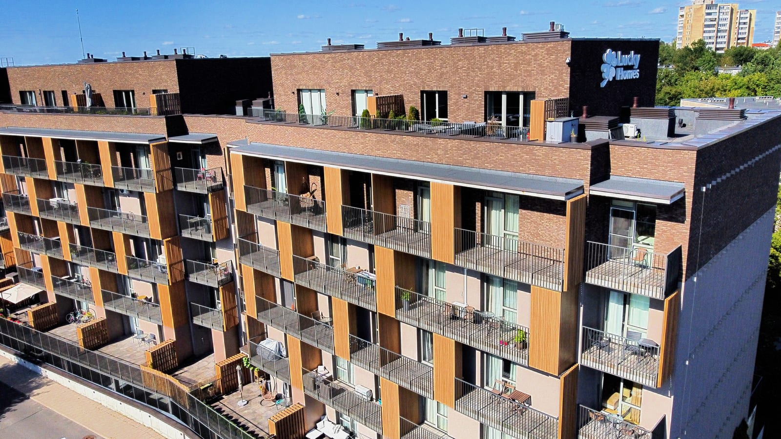 An aerial view along the building with the illuminated Lucky Homes sign fixed to the dark gable end, above balconies set between brick piers and timber-slatted panels.